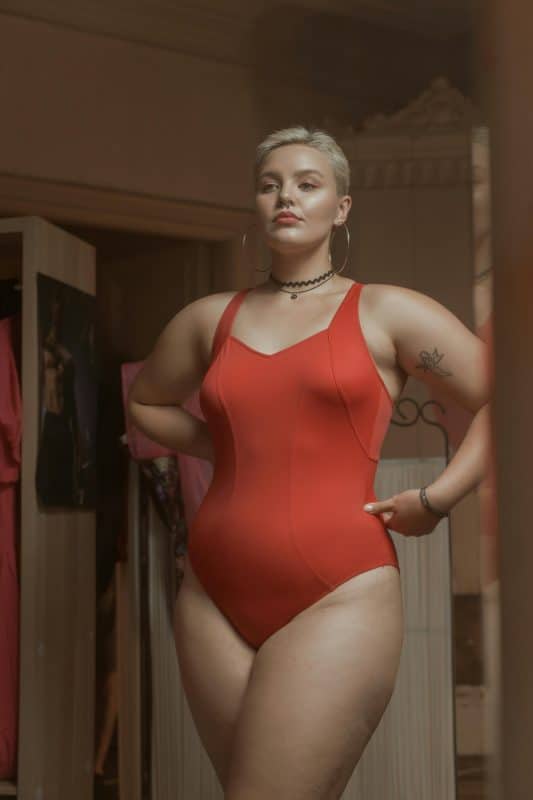 Femme ronde, en maillot rouge, se regardant fièrement dans un miroir. Elle accepte ses formes et ses rondeurs, loin des canons de beauté traditionnels. C'est la base de la sexulité positive