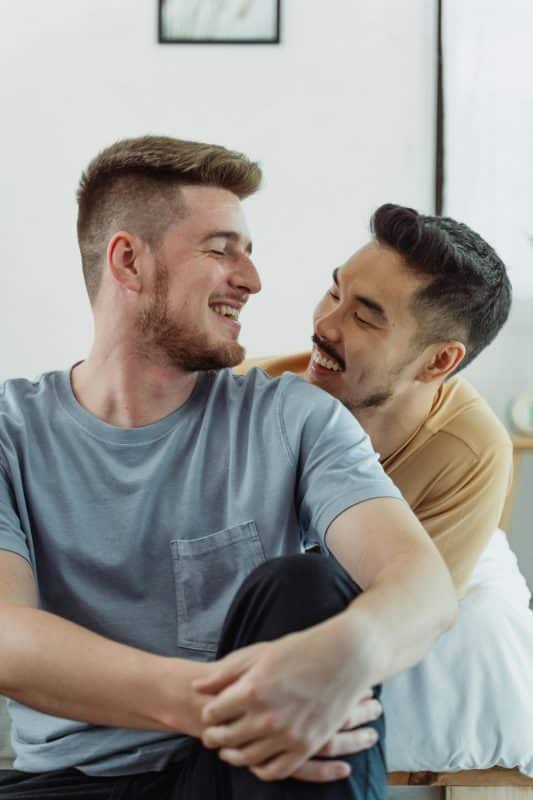 Couple d'hommes rigolant, assis sur leur lit. Parler de ses besoins et de ses envies pour aboutir à une sexualité positive.