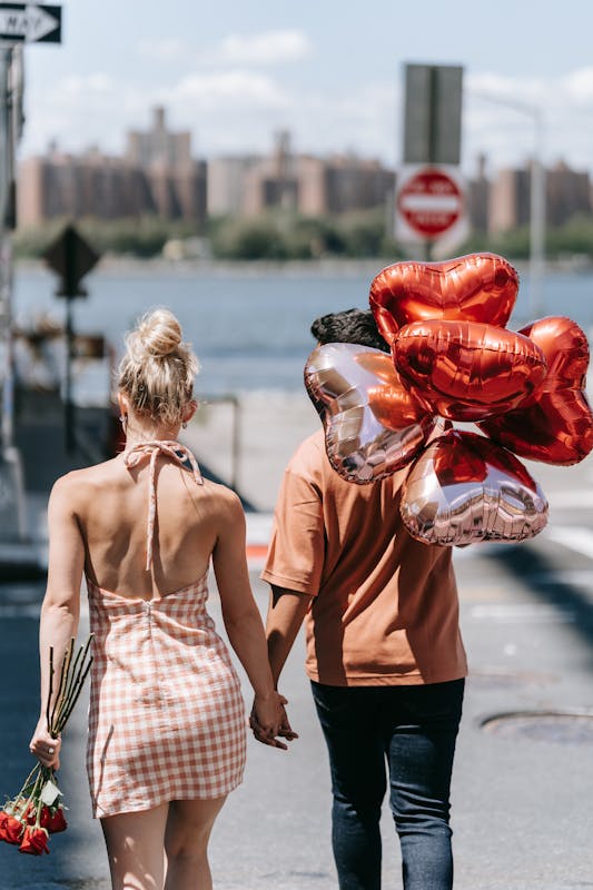 A couple holding hands with heart-shaped balloons and roses, walking in an urban setting. Idée Saint Valentin